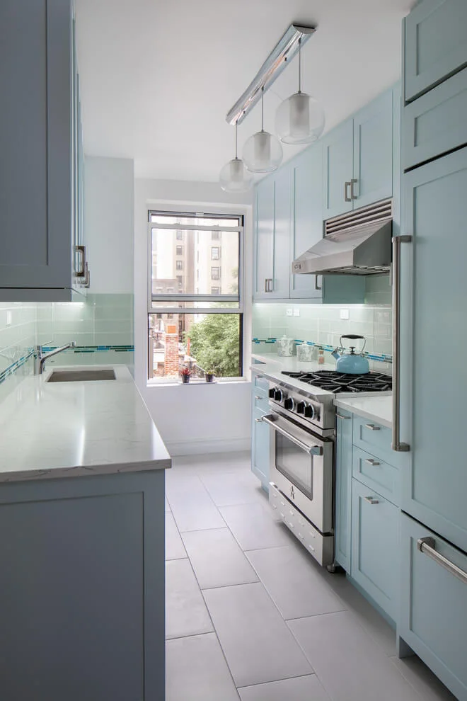 Galley kitchen in a renovated Manhattan prewar apartment with pale blue cabinetry and a window at the end, by Grandeur Hills Group